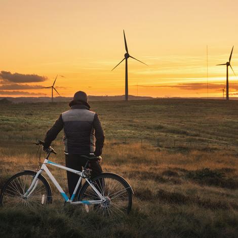A man leaning against a bicycle looks into the distance at the sunset behind wind turbines